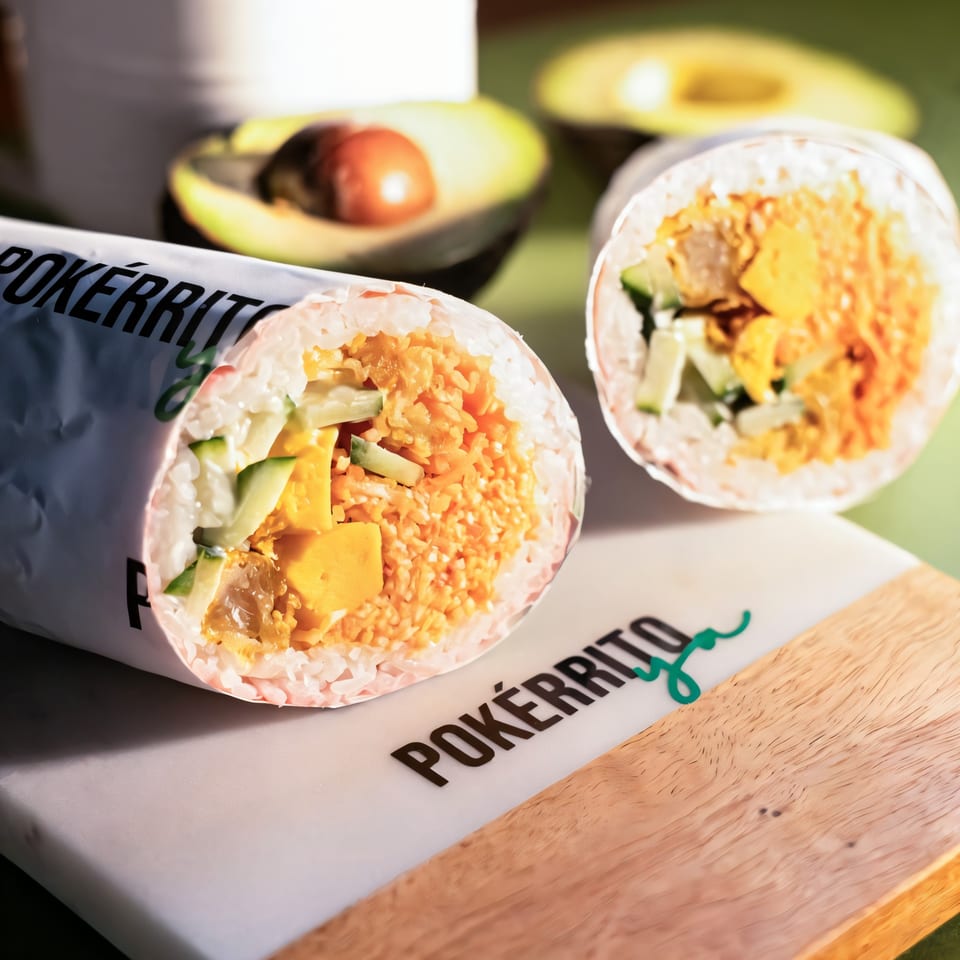 Two Pokérrito Ya poke burritos with avocado halves in the background on a green surface.