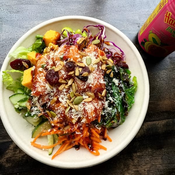 A colorful Pokérrito Ya poke bowl with salmon, vegetables, seeds, and a pink drink can.