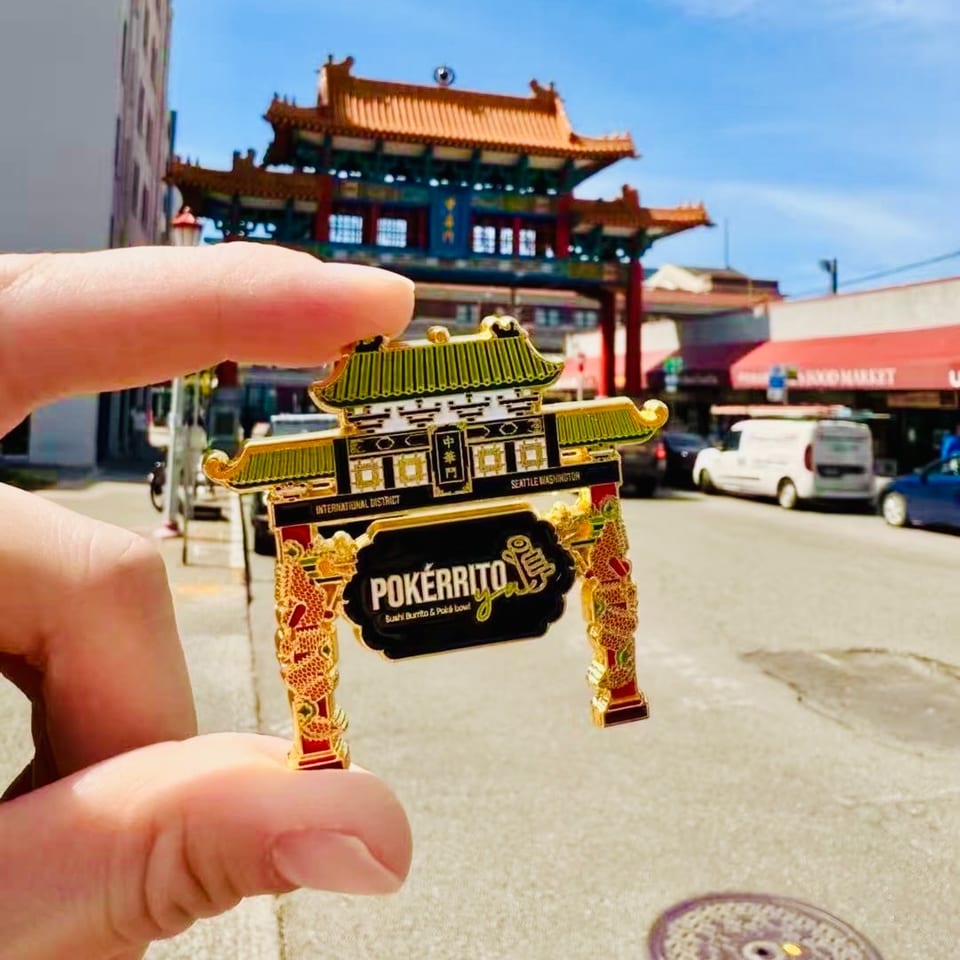 A hand holds a Pokérrito Ya pin in front of the Seattle Chinatown International District gate.