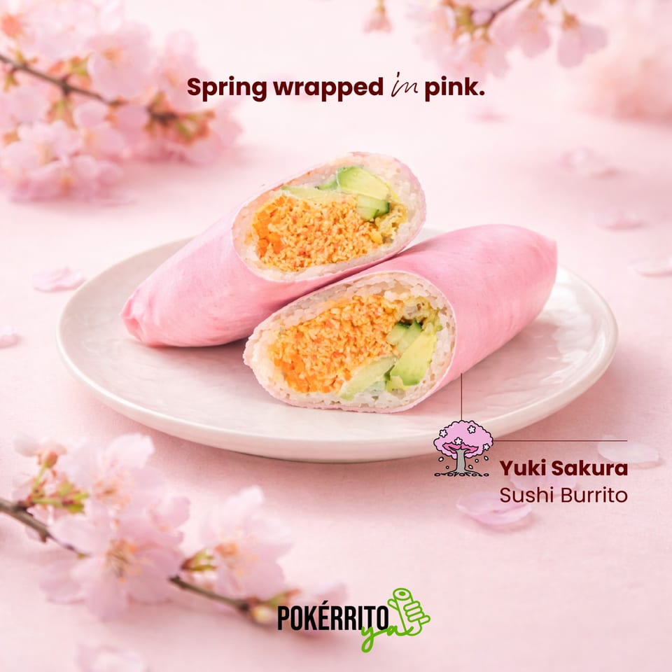 Two pink sushi burritos with avocado and orange filling on a white plate, surrounded by cherry blossoms.