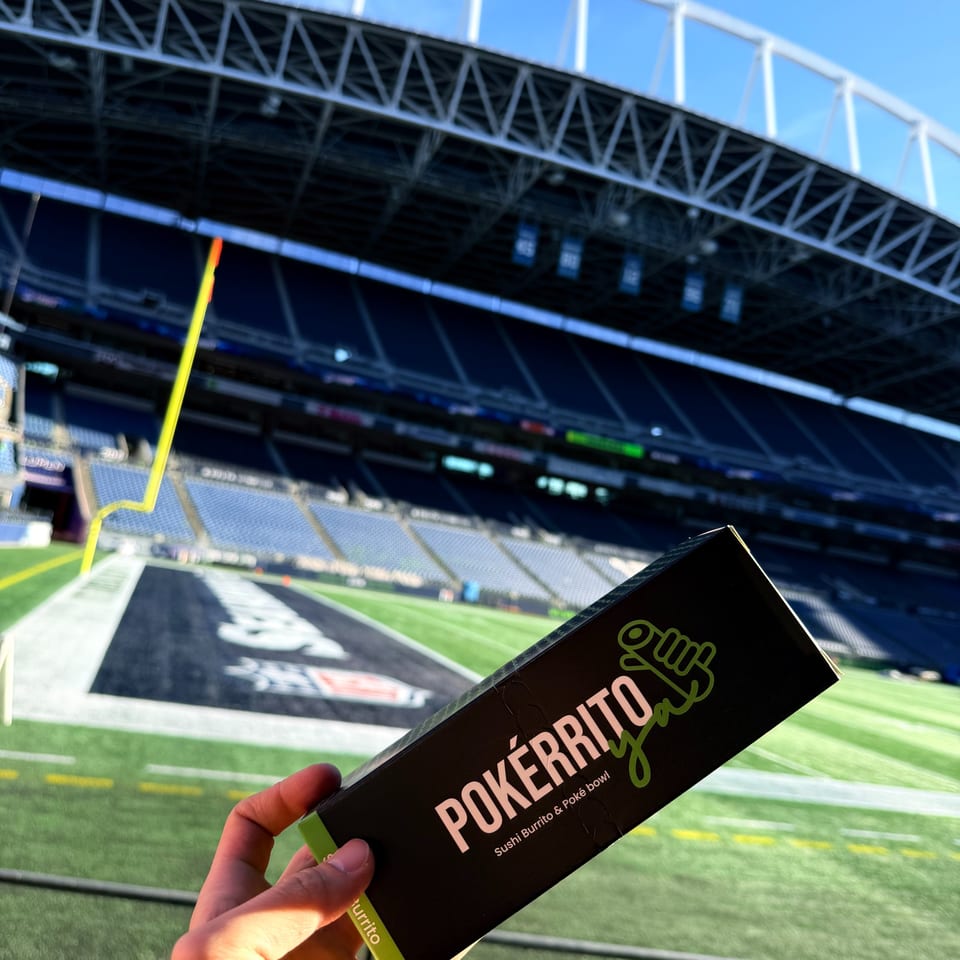 A hand holds a Pokérrito Ya box with a football stadium and field in the background.