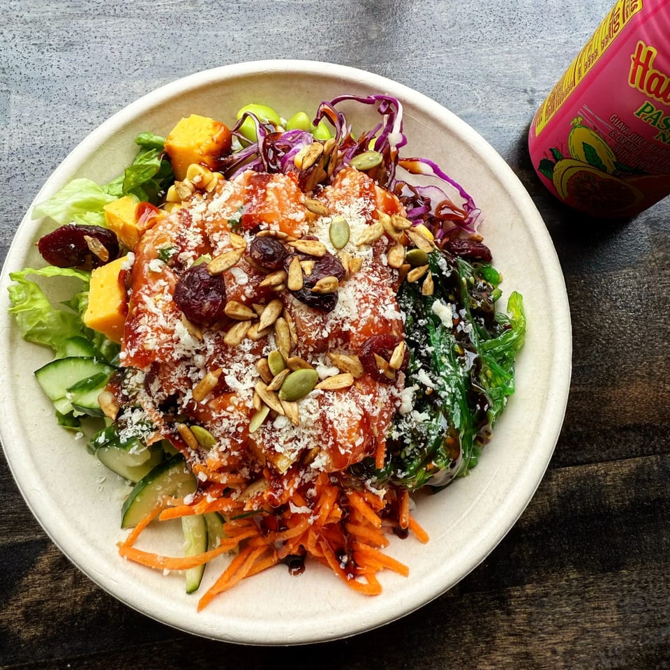 A colorful Pokérrito Ya poke bowl with salmon, vegetables, seeds, and sauce, next to a pink drink.